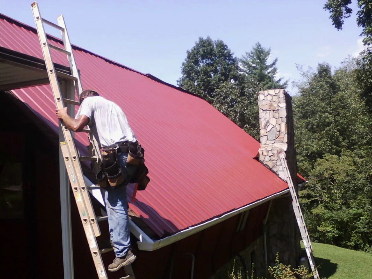Roofer on a ladder beside a red metal roof and stone chimney — Soffit & Fascia Repair by Roofing Brothers in Rolling Meadows